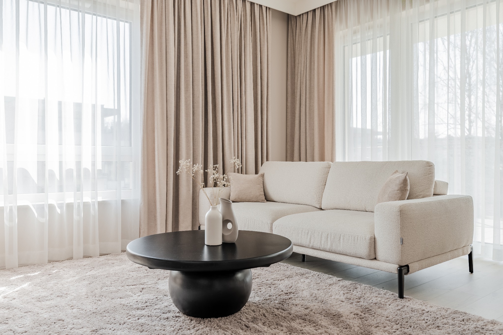 Jasne wnętrze salonu z beżową sofą, ciemnym stolikiem kawowym i puszystym dywanem. Okna z delikatnymi, białymi firanami i beżowymi zasłonami w tle.