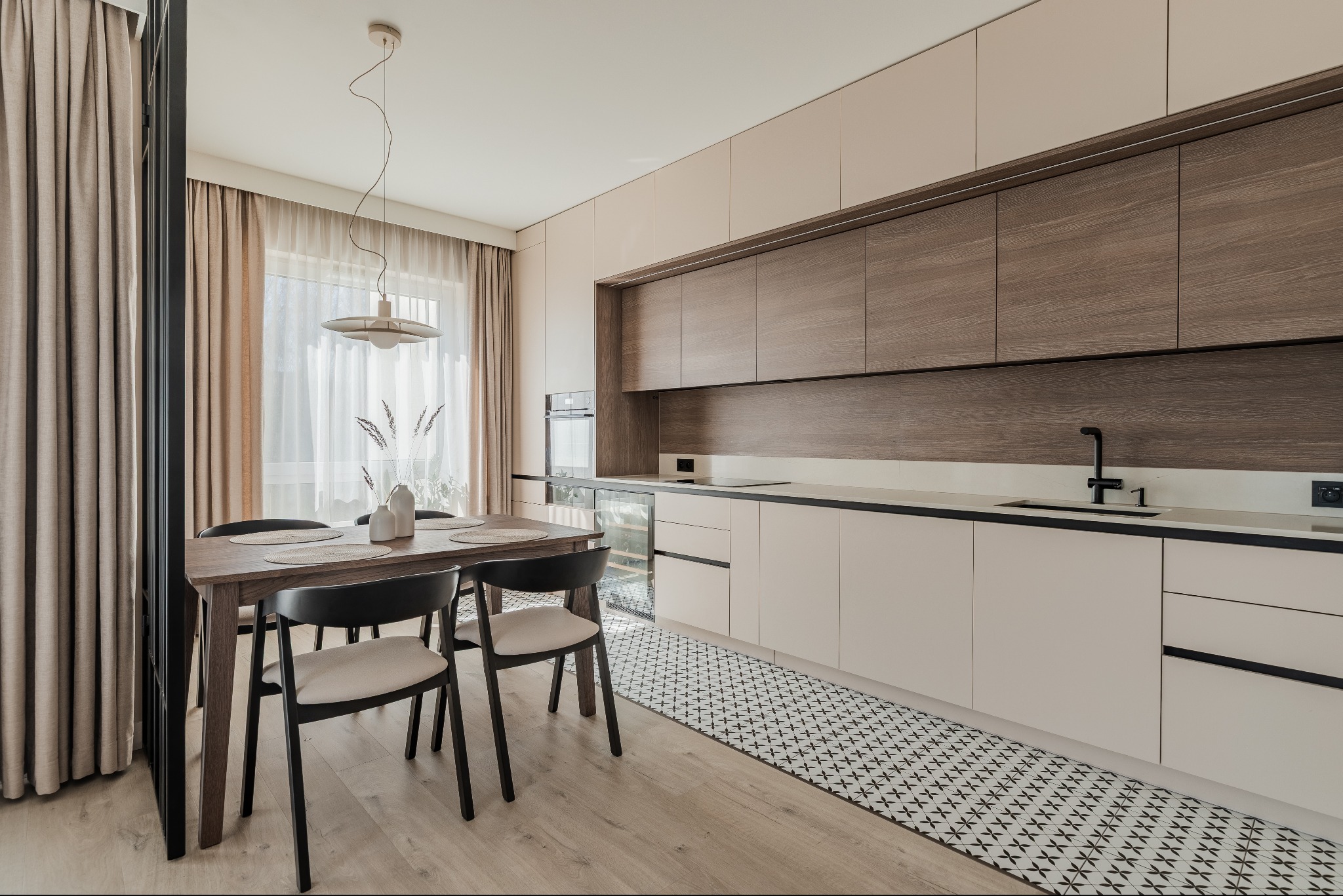 Nowoczesna kuchnia z drewnianymi szafkami i stołem jadalnym. Uwagę przykuwa wzorzysty dywanik i  minimalistyczna lampa nad stołem. Jasne, naturalne światło.