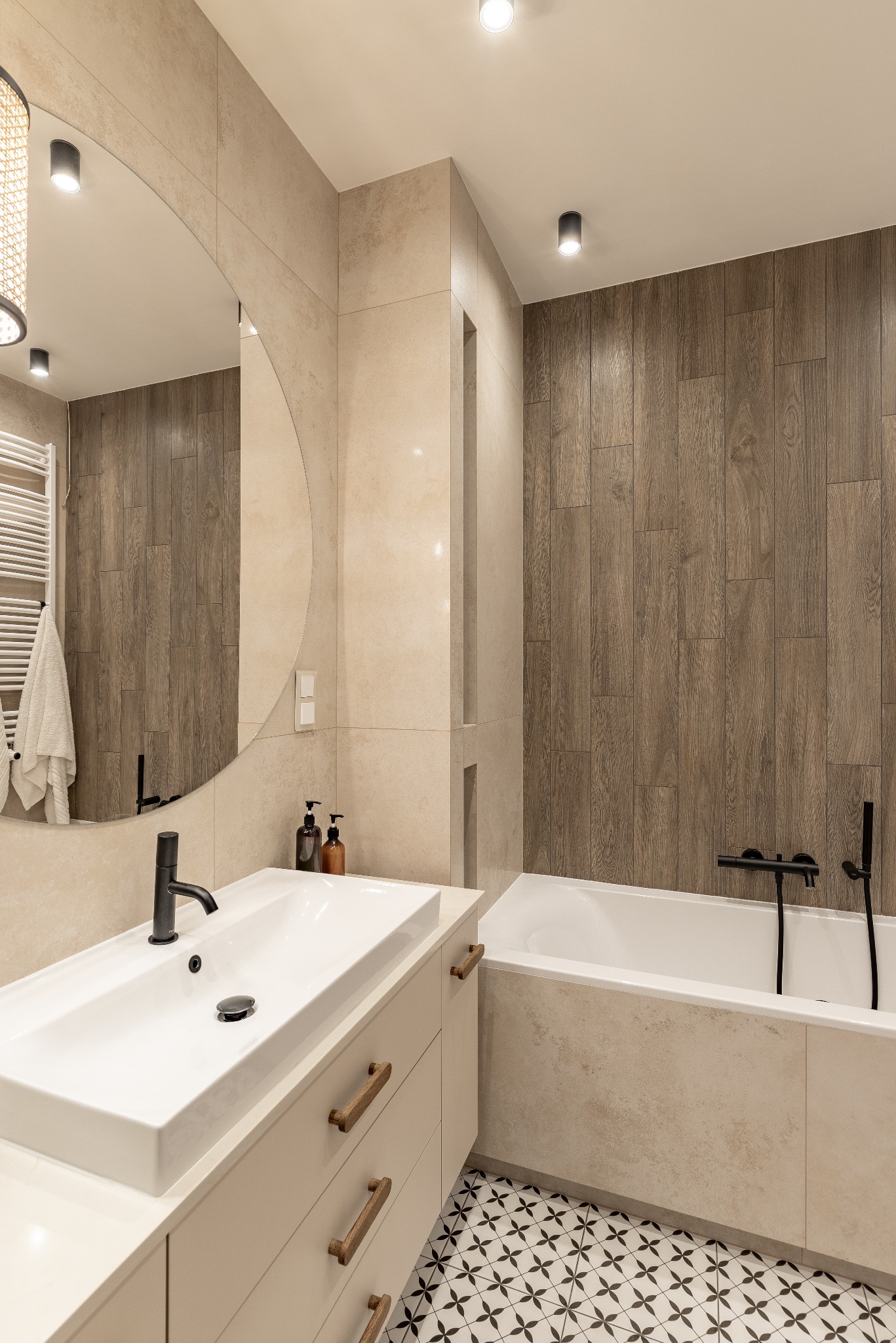 Nowoczesna łazienka z okrągłym lustrem, białą umywalką, drewnianymi akcentami i mozaikową podłogą. Drewnopodobne panele za wanną dodają ciepła.