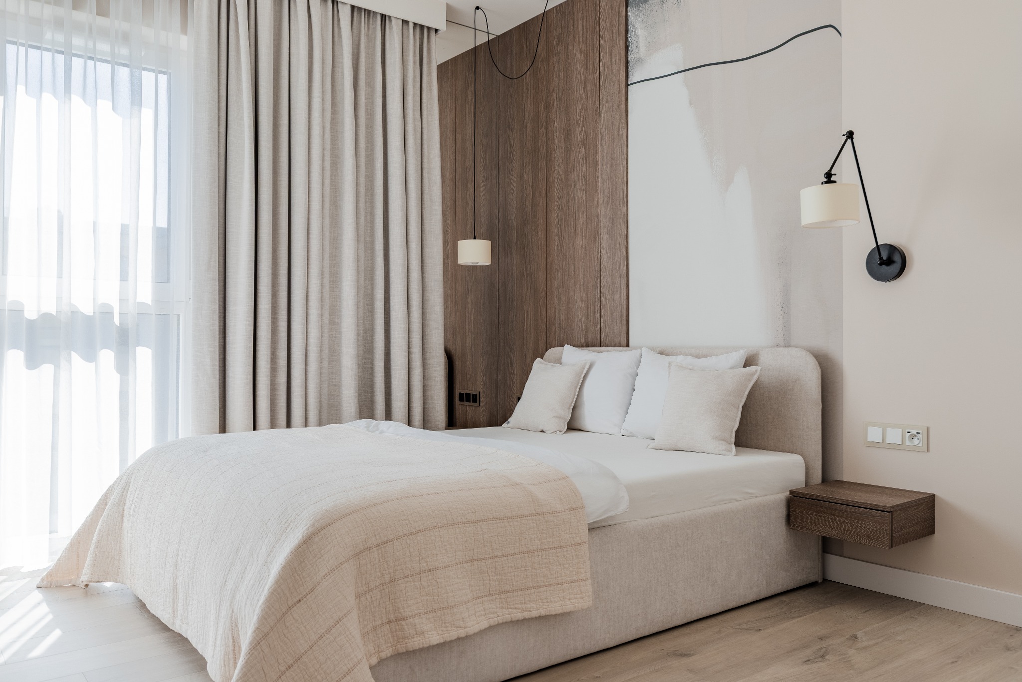 Jasna sypialnia z beżowym łóżkiem, drewnianą ścianą i lampami. Stonowana kolorystyka, naturalne światło wpadające przez okno z zasłonami. Minimalistyczny design.