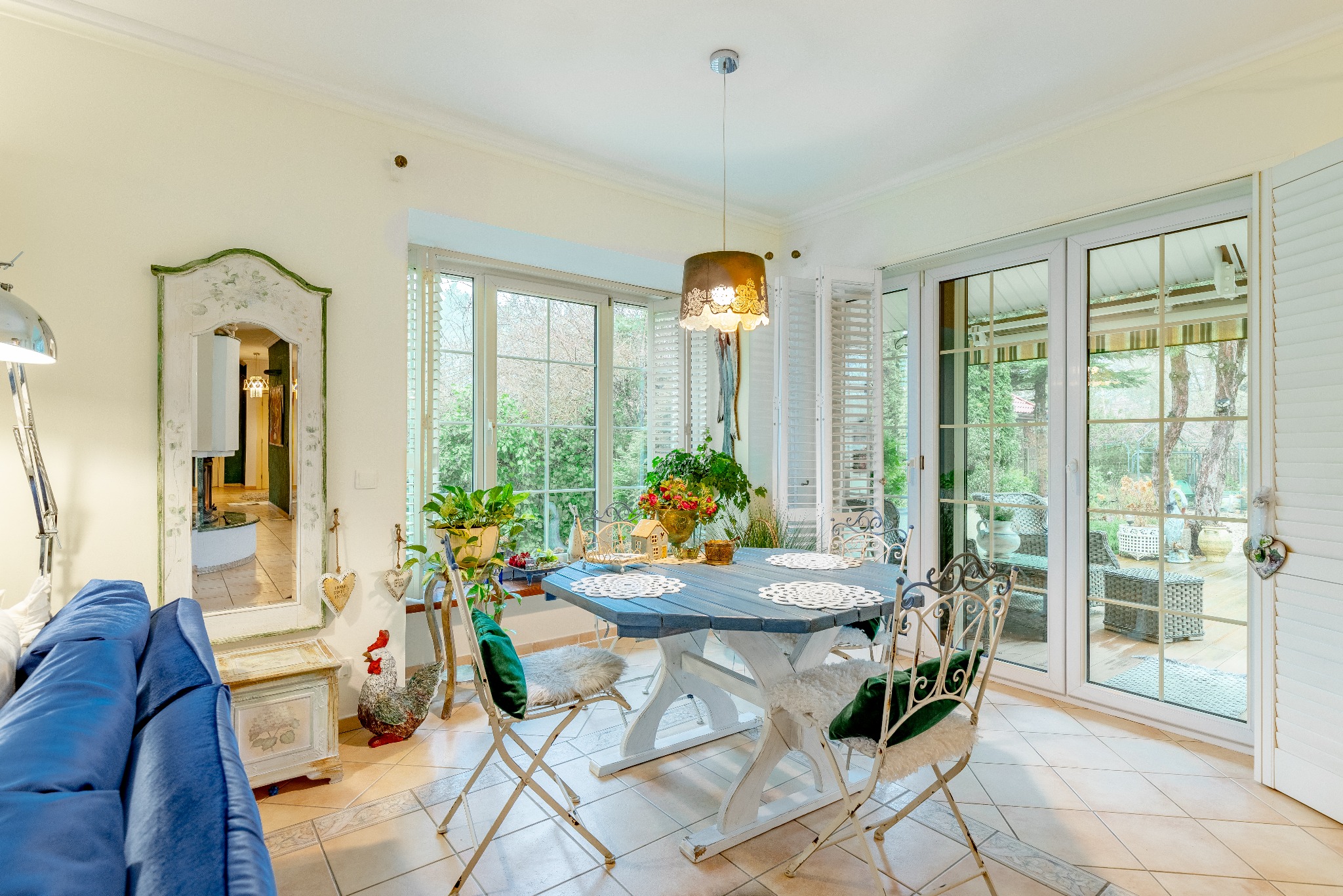 Jasne wnętrze jadalni z niebieskim stołem, białymi krzesłami i dużymi oknami z widokiem na ogród. Dekoracyjny wystrój, lustro i lampa wisząca dodają uroku.