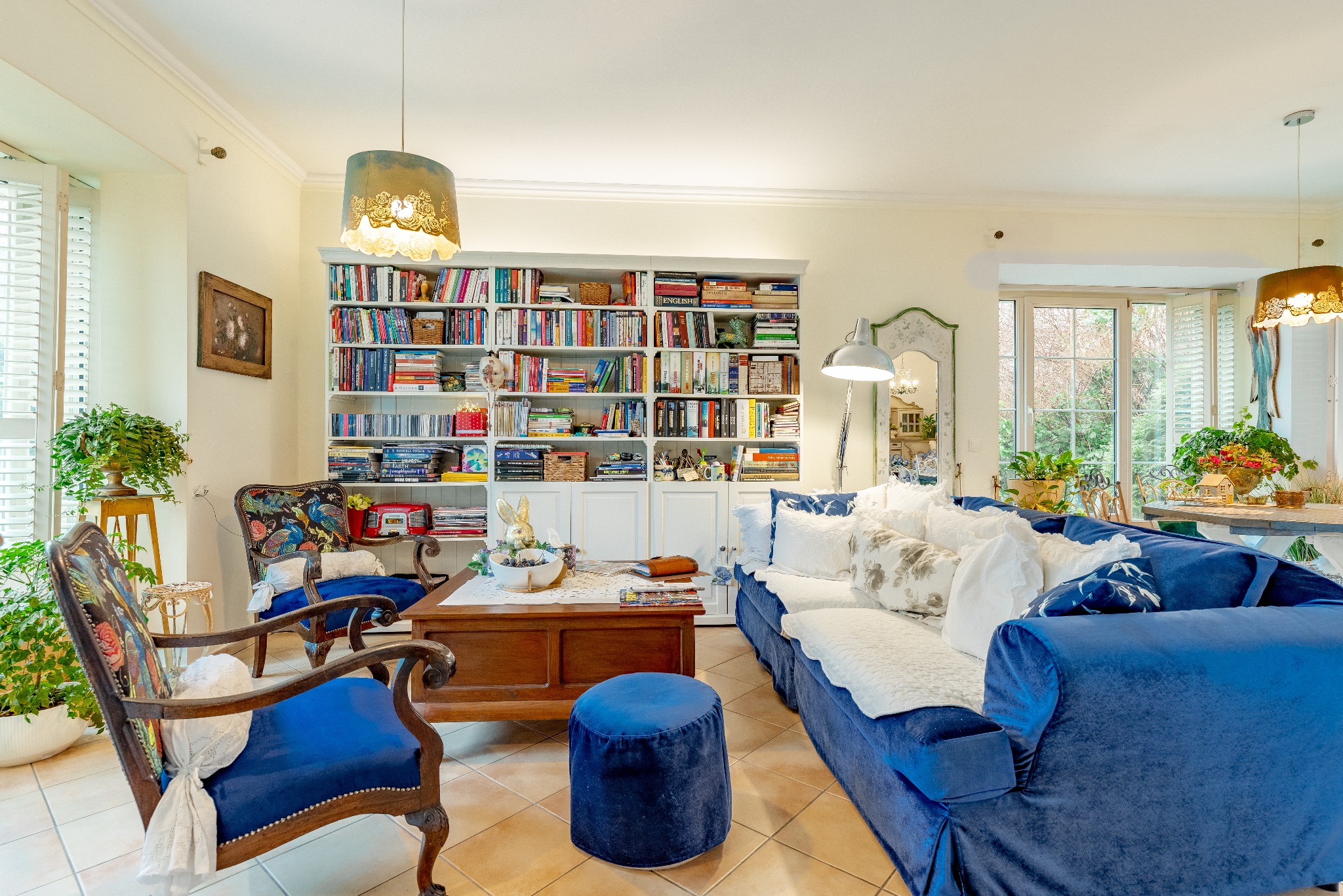 Salon z biblioteczką, niebieską sofą i fotelami w stylu vintage. Jasne wnętrze z roślinami i dekoracyjnymi lampami. Fotografia wnętrza nieruchomości.
