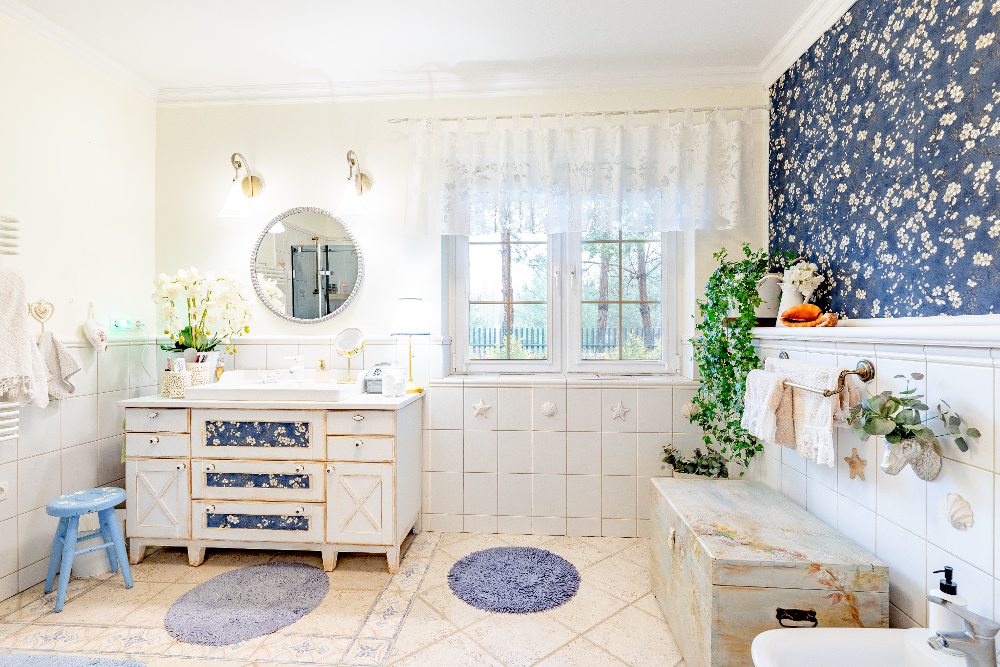 Jasna łazienka z białymi kaflami, szafką z kwiatowym dekorem i okrągłym lustrem. Dekoracyjne detale: morskie gwiazdy, muszle, niebieska kwiecista tapeta.