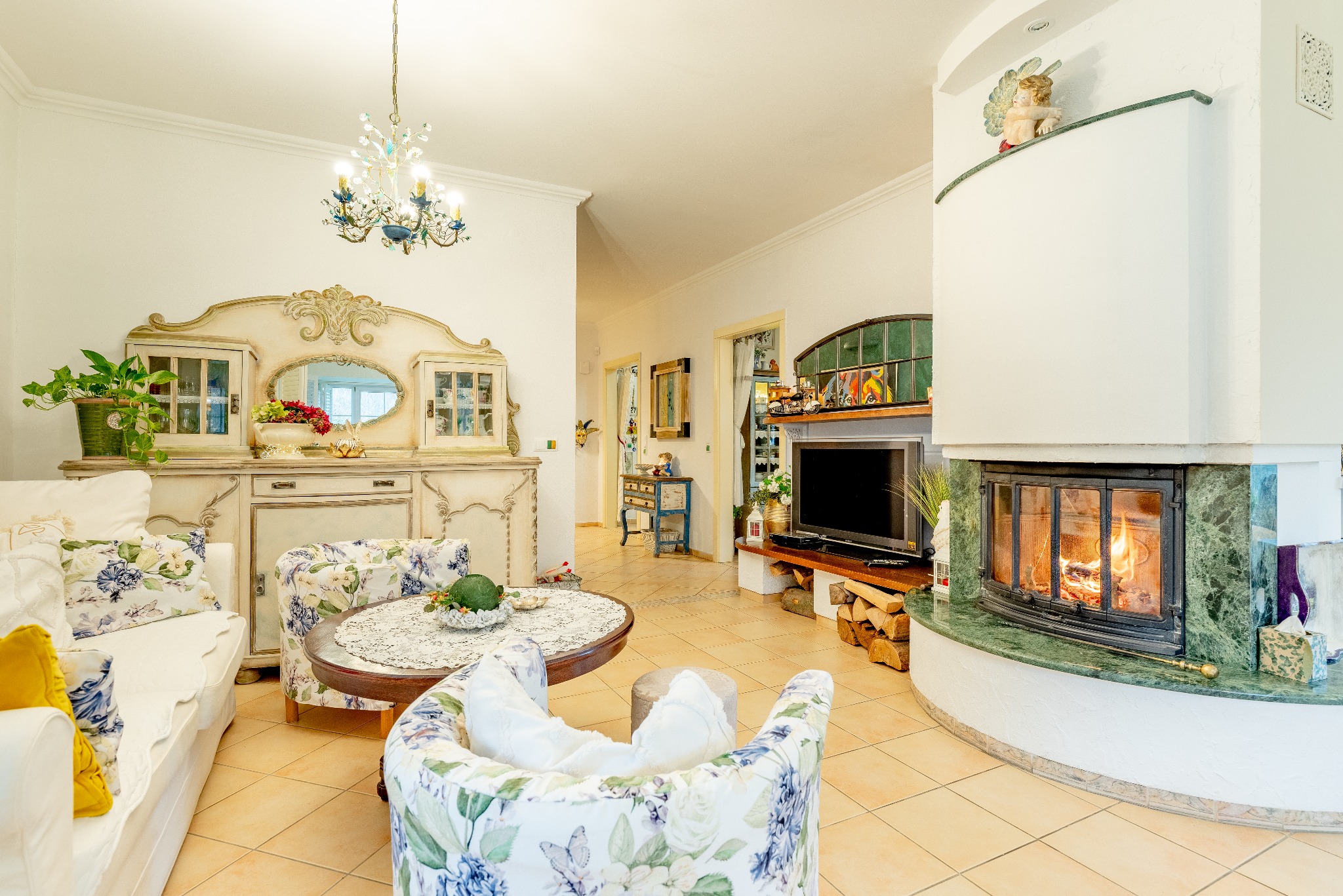 Jasny salon z kominkiem obudowanym zielonym marmurem, antycznym kredensem i kwiatowymi fotelami. Na stole serweta, a w kominku płonie ogień.