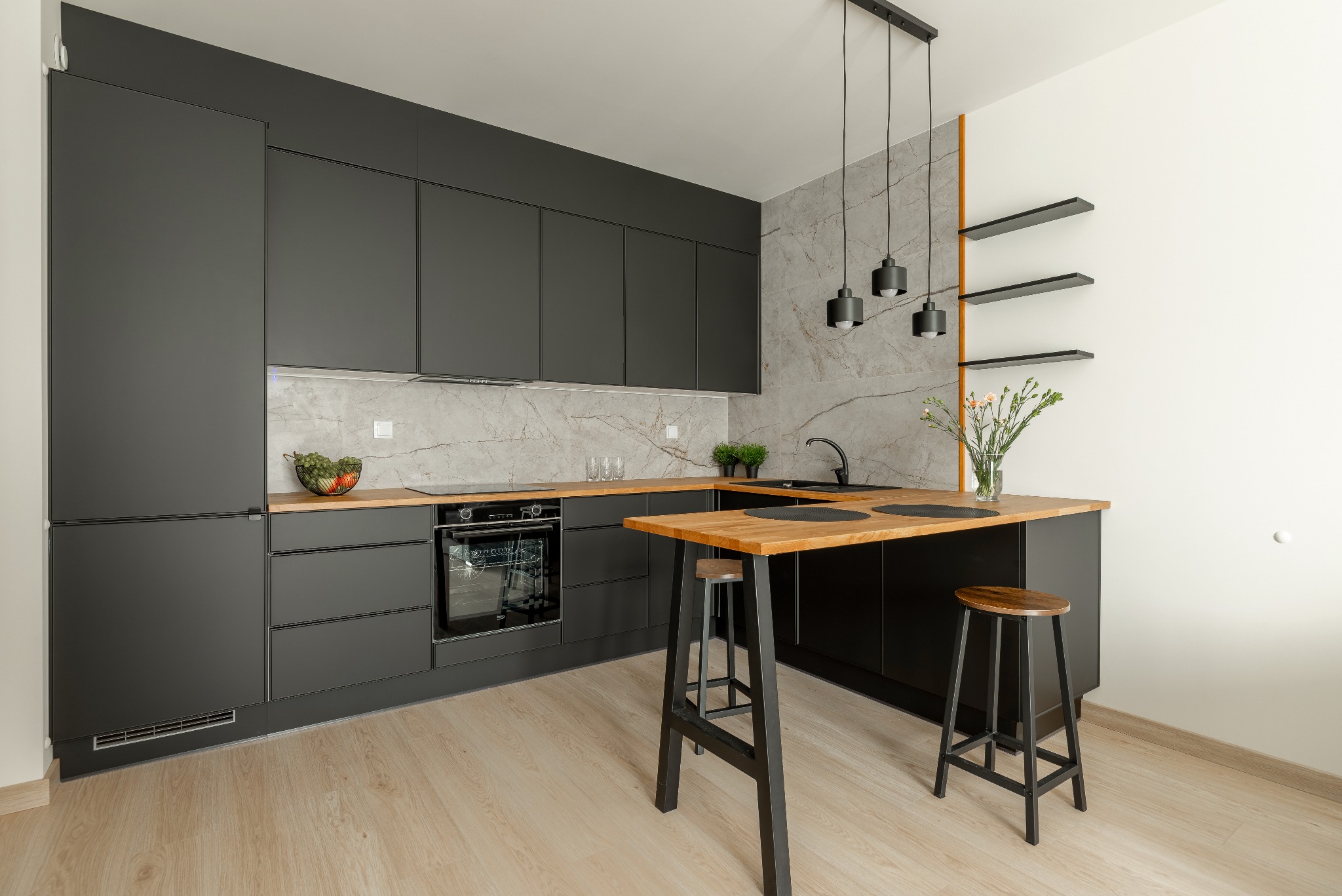 Nowoczesna, czarna kuchnia z drewnianymi blatami i marmurowym backsplashiem. Dwa stołki barowe stoją przy wyspie kuchennej. Minimalistyczne oświetlenie i kwiaty dodają charakteru.