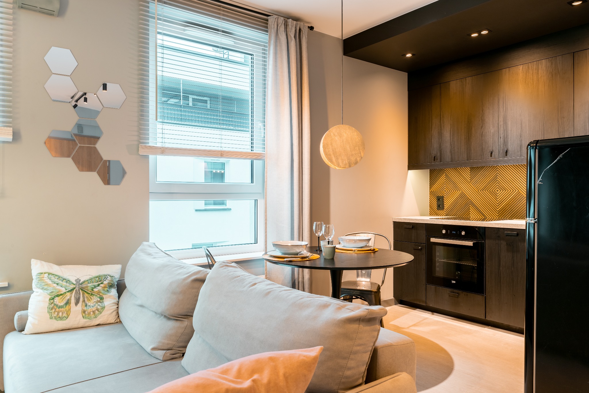 Jasne wnętrze mieszkania z aneksem kuchennym i okrągłym stołem. Dekoracyjne lustra na ścianie. Poduszka z motylem na sofie. Nowoczesny wystrój.