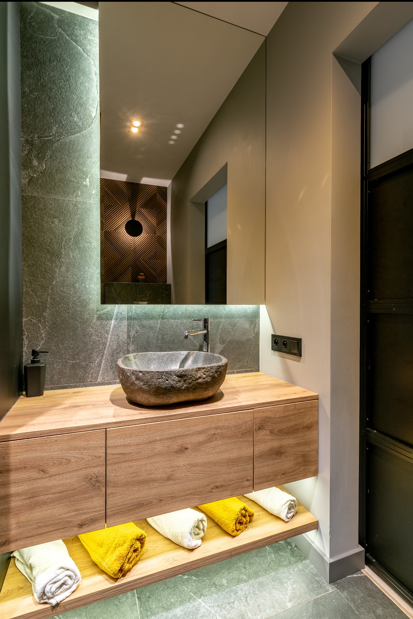 Nowoczesna łazienka: kamienna umywalka na drewnianej szafce z oświetleniem LED, lustro z odbiciem dekoracyjnej ściany. Szare płytki i akcenty czerni.