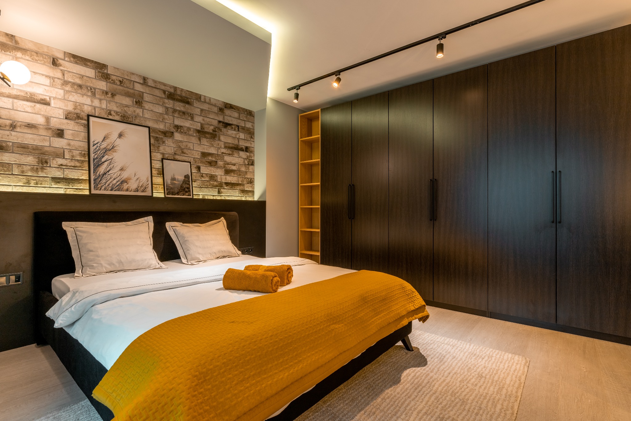 Nowoczesna sypialnia z ceglaną ścianą, łóżkiem z żółtą narzutą i ciemną szafą wnękową. Oświetlenie punktowe i obrazy na ścianie dodają wnętrzu charakteru.