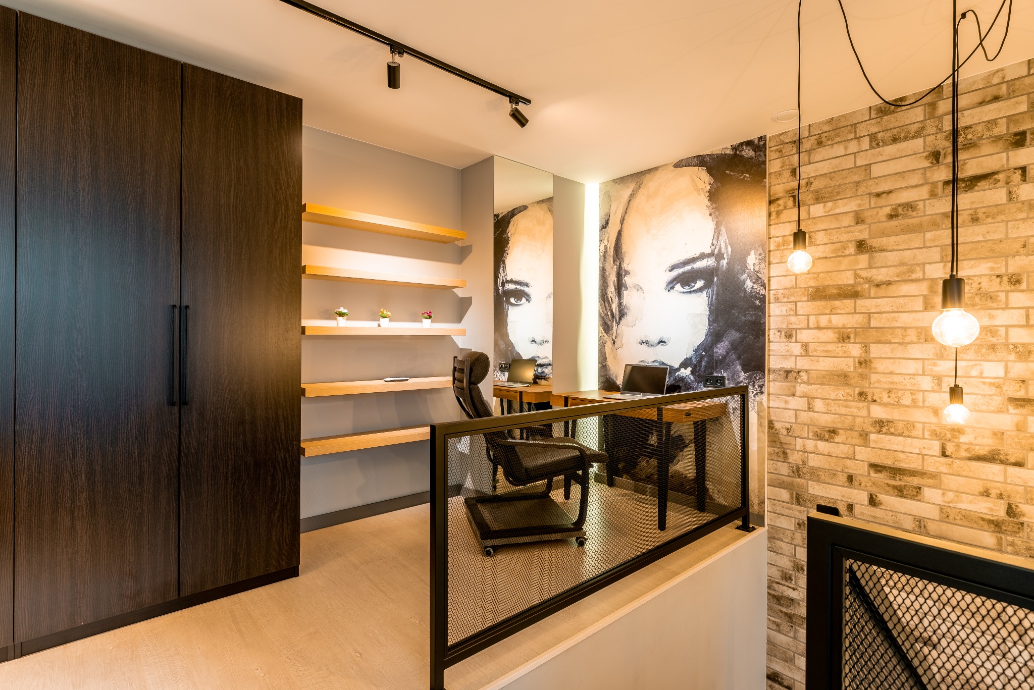 Nowoczesne biuro domowe z ceglaną ścianą, minimalistycznym biurkiem i regałami. Duże okno wpuszcza naturalne światło. Wyjątkowa tapeta z motywem kobiecej twarzy.