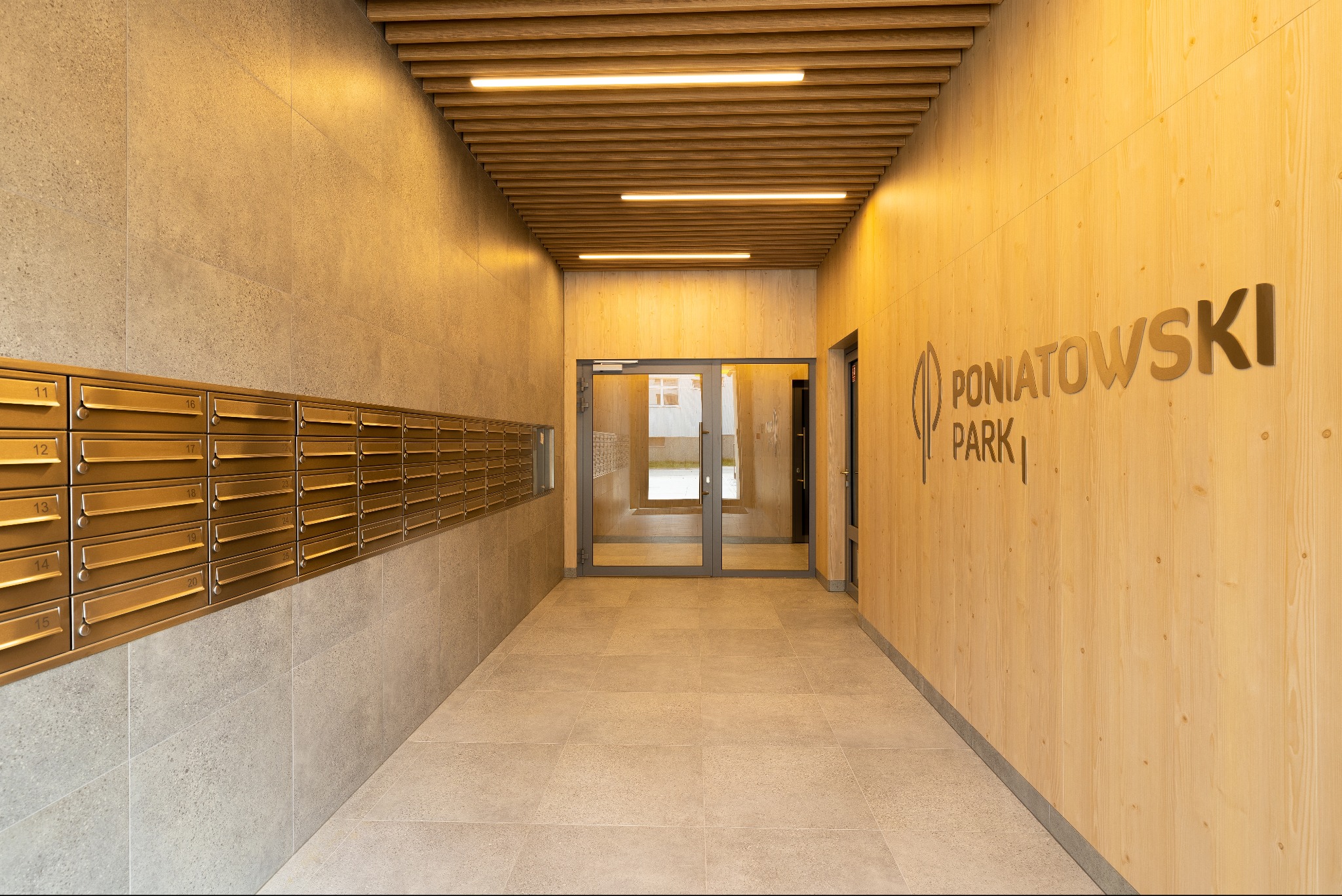 Jasne, nowoczesne wnętrze holu wejściowego z drewnianą boazerią i rzędami skrzynek pocztowych. Widoczne logo 'Poniatowski Park' i przeszklone drzwi.