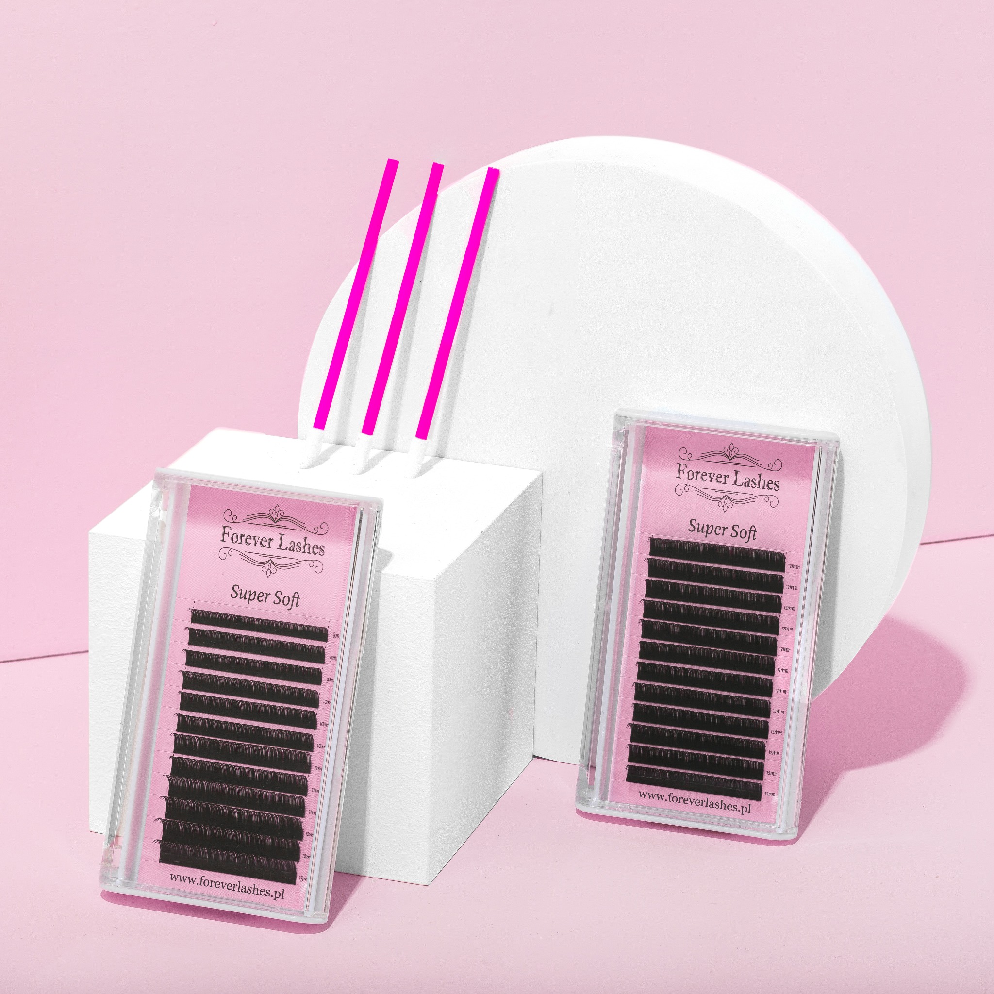 Dwa opakowania rzęs 'Forever Lashes Super Soft' na białych geometrycznych blokach i różowym tle, z trzema aplikatorami. Fotografia produktowa.