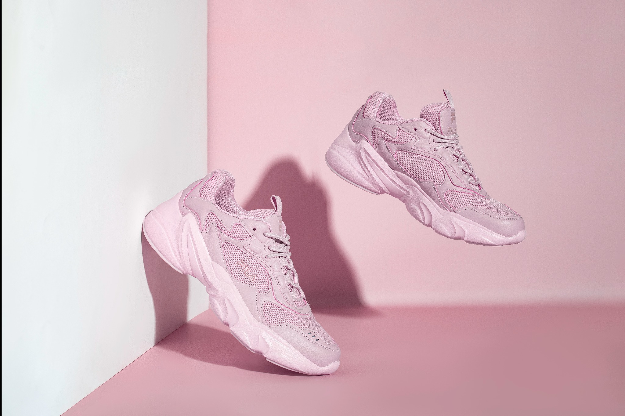 Stylizowana fotografia produktowa: dwa identyczne, różowe sneakersy unoszą się w przestrzeni na tle różowo-białego, minimalistycznego tła. Cienie dodają głębi.