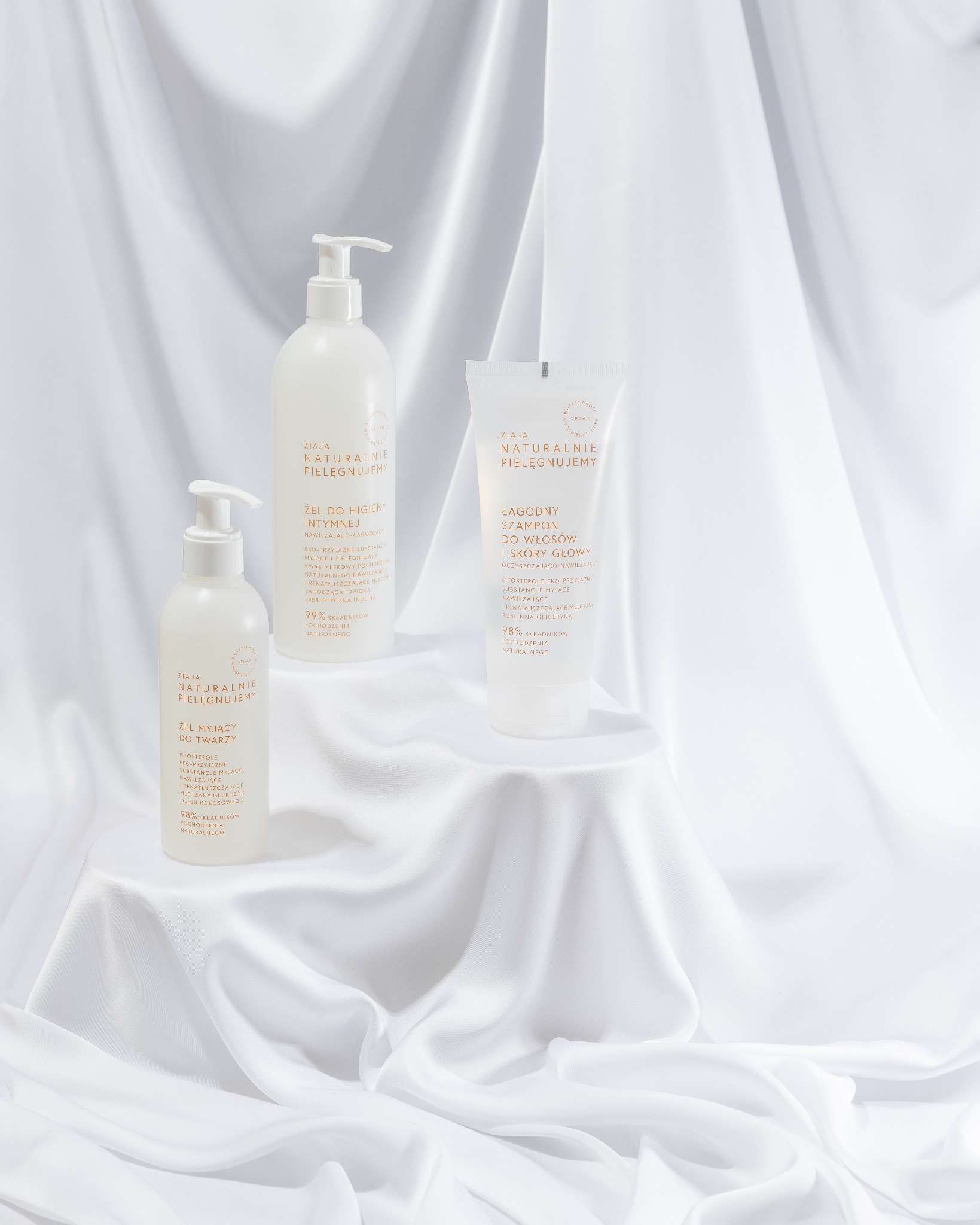 Stylizowana fotografia produktowa: Trzy białe kosmetyki Ziaja Naturalnie Pielęgnujemy na tle białej, połyskującej tkaniny. Żel do higieny intymnej, żel do twarzy i szampon.