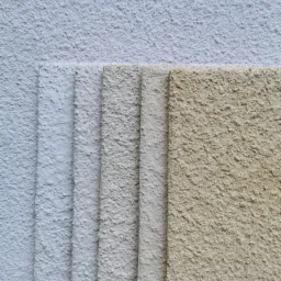 Próbki tynków elewacyjnych o różnej kolorystyce i fakturze, ułożone warstwami na tle ściany z nałożonym jasnym tynkiem.