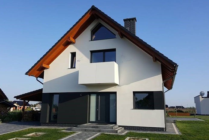 Nowoczesny dom jednorodzinny z jasną elewacją, ciemnymi oknami i drewnianym wykończeniem dachu, widziany z niskiej perspektywy w słoneczny dzień.