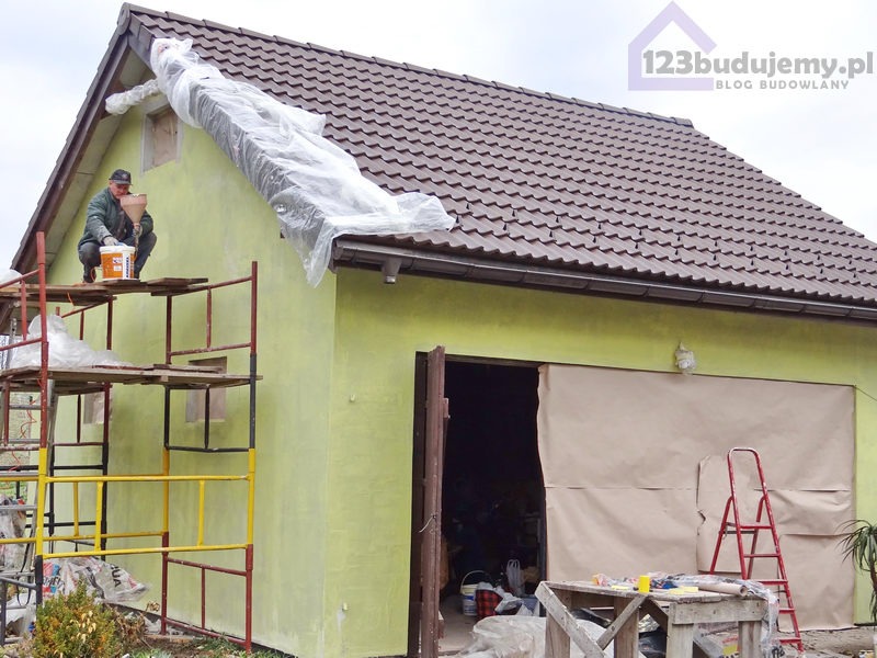 Pracownik na rusztowaniu w trakcie malowania elewacji budynku, zabezpieczone okno folią, przygotowane wejście do garażu, widoczny napis '123budujemy.pl' w prawym górnym rogu.