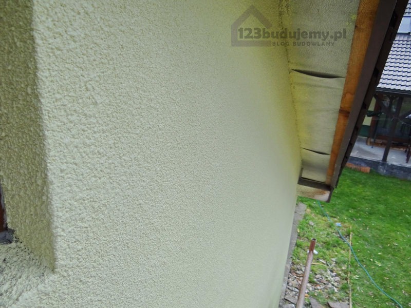 Ściana budynku po nałożeniu struktury tynku baranka w jasnym odcieniu, widoczne elementy dachu i fragment ogrodu. Na ścianie widoczny znak wodny strony internetowej.