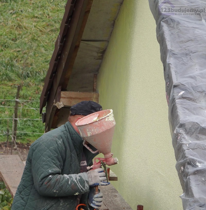 Pracownik w zielonej kurtce aplikuje tynk strukturalny pistoletem natryskowym na elewację budynku, widoczne rusztowanie i zabezpieczona folią krawędź dachu.