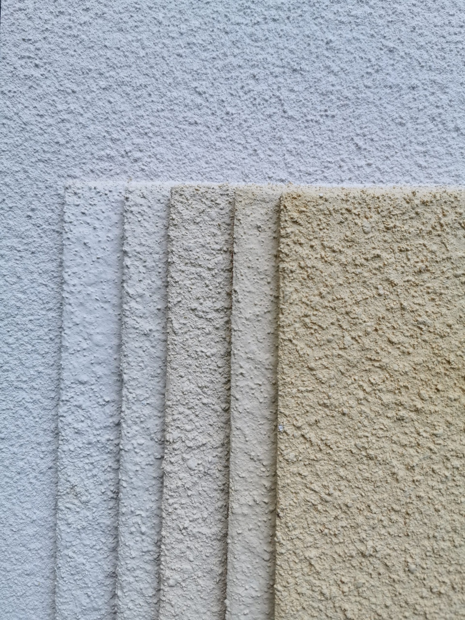 Próbki tynków elewacyjnych o różnej kolorystyce i fakturze, ułożone warstwami na tle ściany z nałożonym jasnym tynkiem.