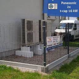 Dwie jednostki zewnętrzne Panasonic t-cap 16 kW zamontowane na betonowych podstawach przy ścianie budynku, widoczne przez metalowe ogrodzenie. Na ogrodzeniu tabliczka informacyjna firmy Virtus Sun,...