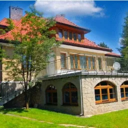 Dwupiętrowy budynek z czerwoną dachówką, balkonem z metalową balustradą i kamienną elewacją parteru z łukowatymi oknami, otoczony zielenią drzew i trawnikiem.