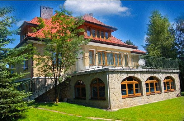 Dwupiętrowy budynek z czerwoną dachówką, balkonem z metalową balustradą i kamienną elewacją parteru z łukowatymi oknami, otoczony zielenią drzew i trawnikiem.