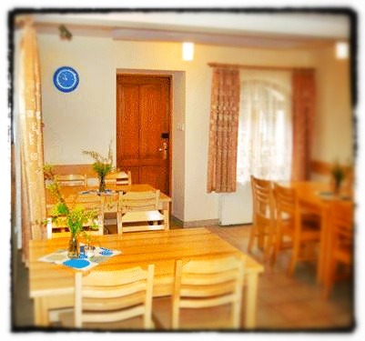 Jasne wnętrze z drewnianymi stołami i krzesłami, na stołach wazony z polnymi kwiatami, na ścianie niebieski zegar.