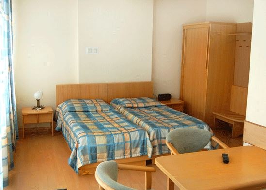 Pokój z dwoma łóżkami pojedynczymi z niebiesko-pomarańczową pościelą w kratę, drewnianym biurkiem z pilotem, szafą i lampką nocną na stoliku.