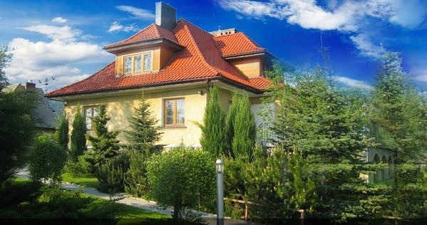 Dwupiętrowy budynek z pomarańczowym dachem, otoczony zielenią i drzewami iglastymi, na tle błękitnego nieba z chmurami.