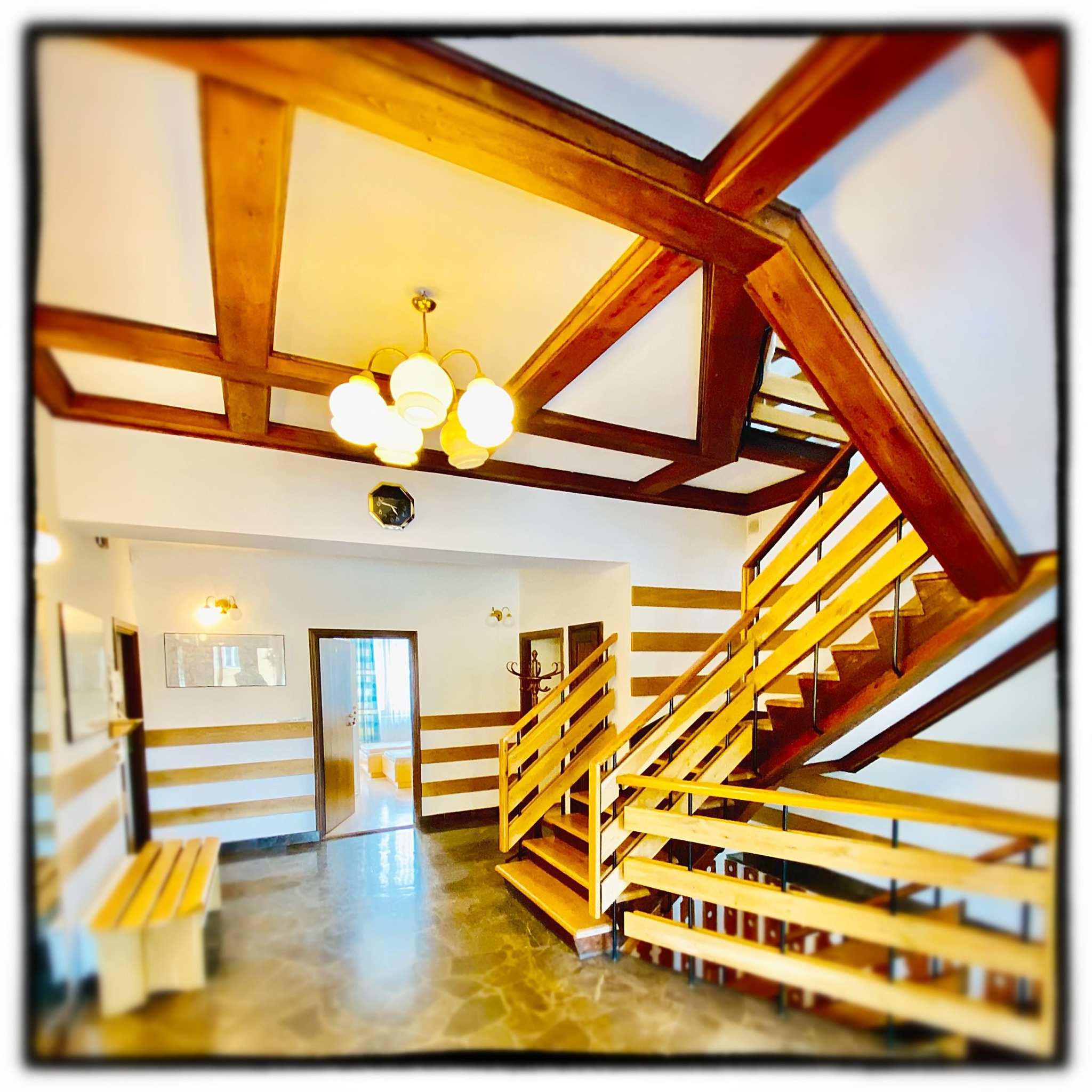 Wnętrze kliniki z drewnianymi schodami, belkami stropowymi i lampą wiszącą, widok z dołu na korytarz z ławką i drzwiami do pokoi.