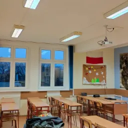 Wnętrze klasy szkolnej z rzędami drewnianych ławek i krzeseł, okna z niebieskimi zasłonami, biała ściana z tablicą korkową i plakatem Twierdzy Tobruk, oświetlenie sufitowe i projektor