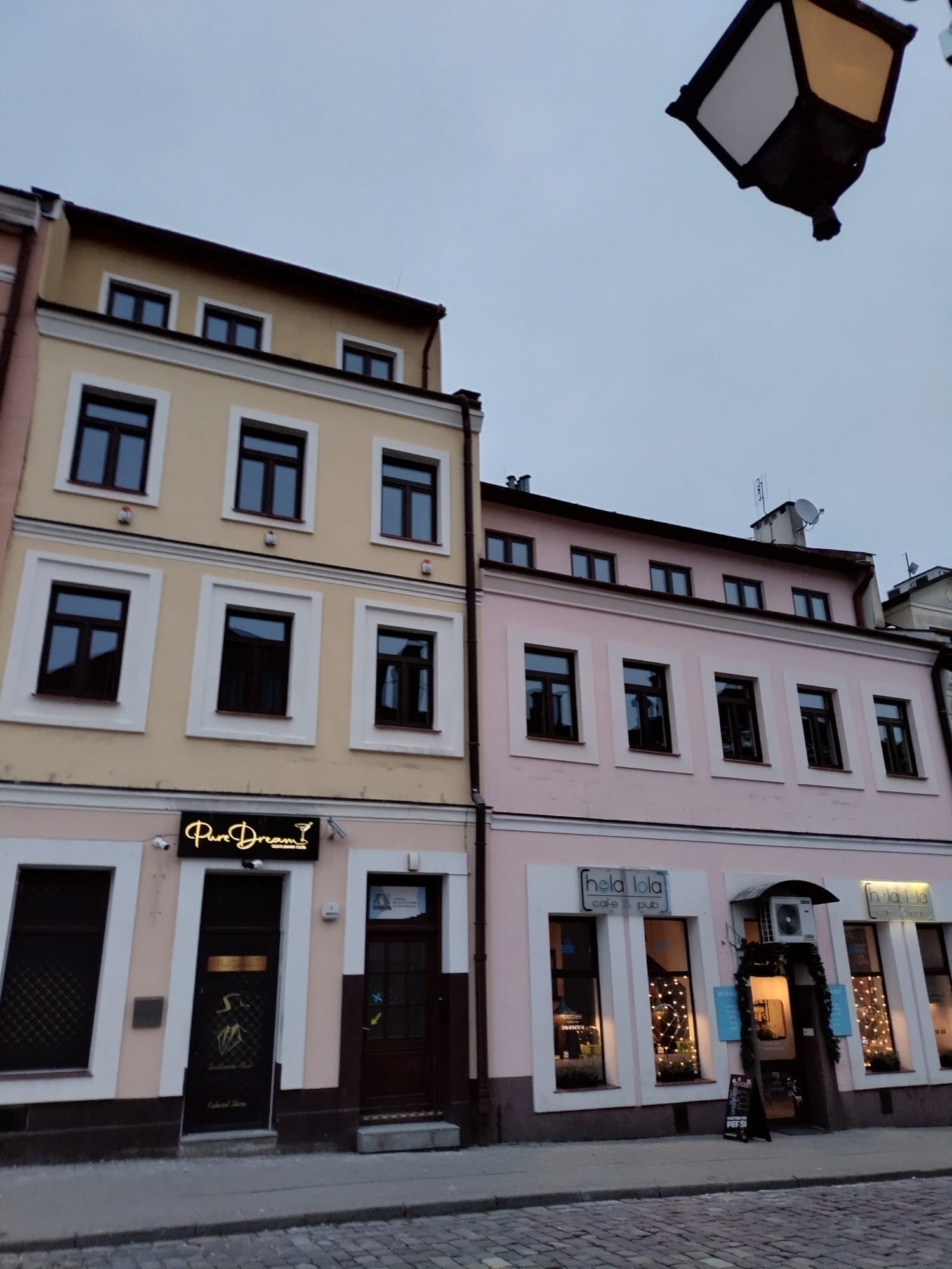 Fasady dwóch kamienic w pastelowych kolorach z ciemnymi oknami, widok na ulicę z kostki brukowej i latarnię uliczną.