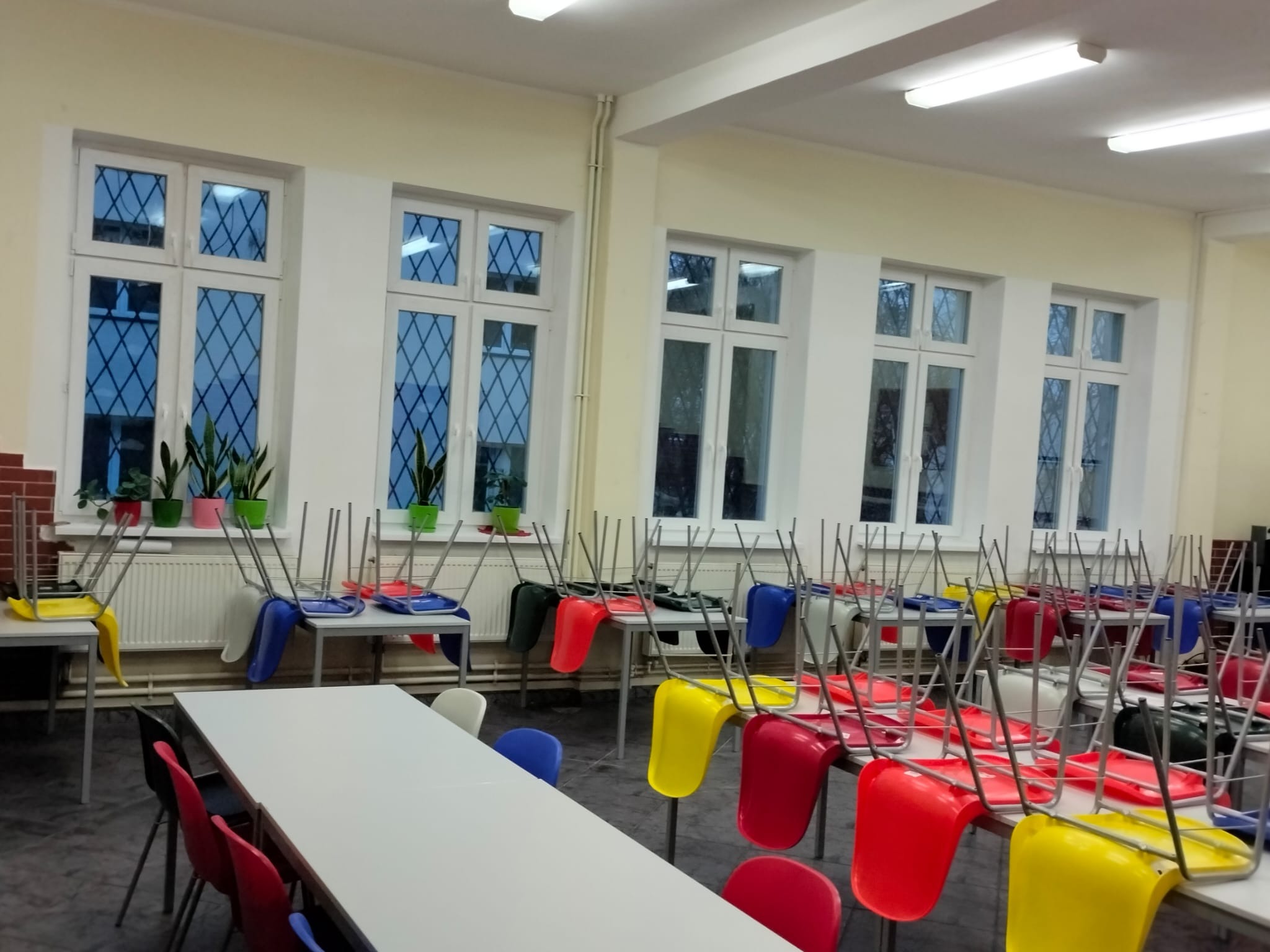 Sala z rzędami stołów i krzeseł ustawionych na blatach, białe okna z kratami i rośliny doniczkowe na parapetach.