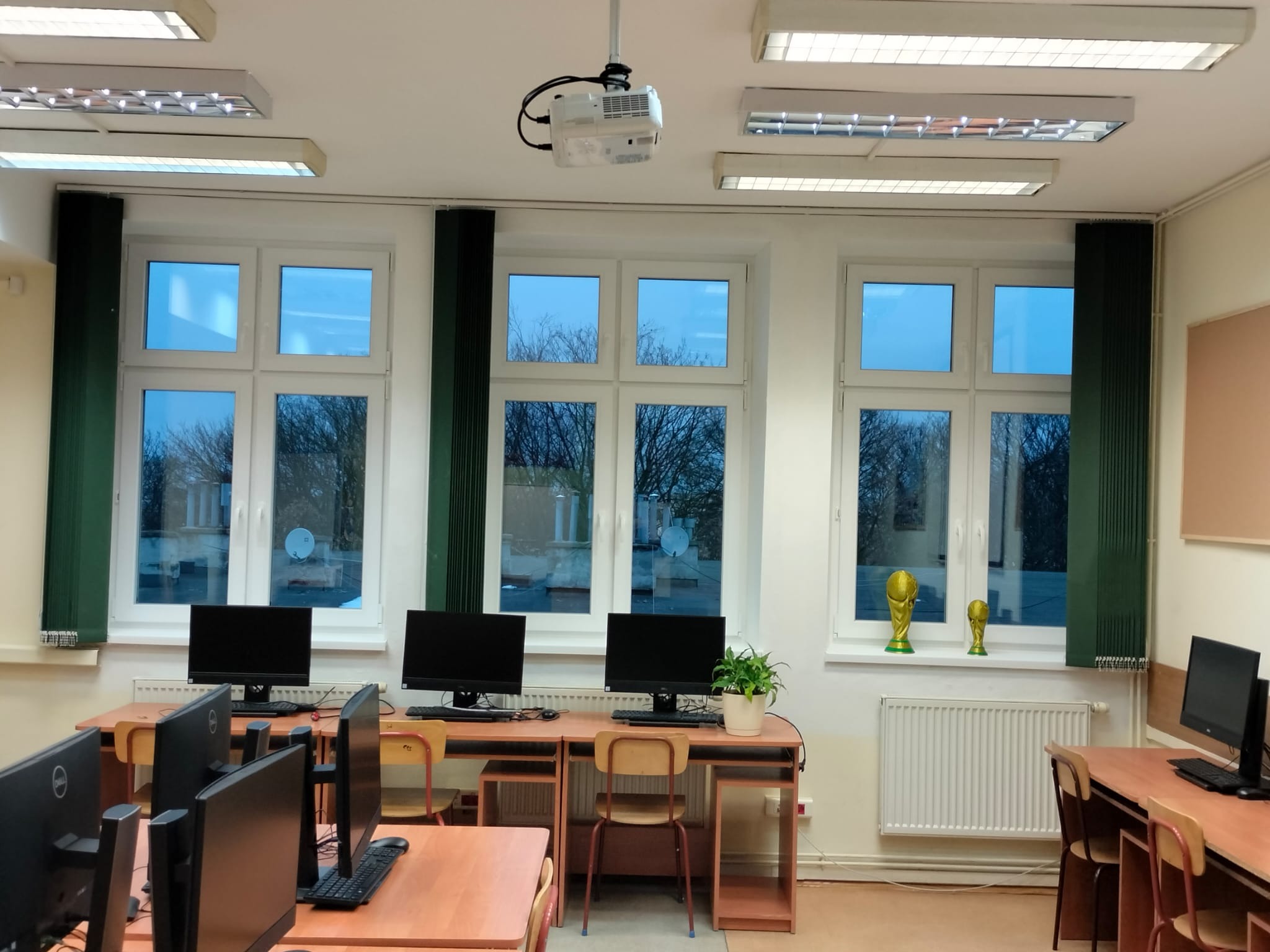 Sala komputerowa z rzędem biurek i komputerów pod oknami z zielonymi roletami; na parapecie stoją dwie złote statuetki, a z sufitu zwisa projektor.