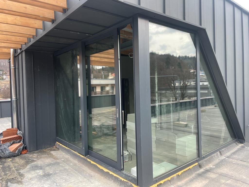 Nowoczesne okno przesuwne z szarymi ramami, zamontowane w skośnej ścianie budynku z metalową elewacją, widok z zewnątrz na taras z drewnianą pergolą.