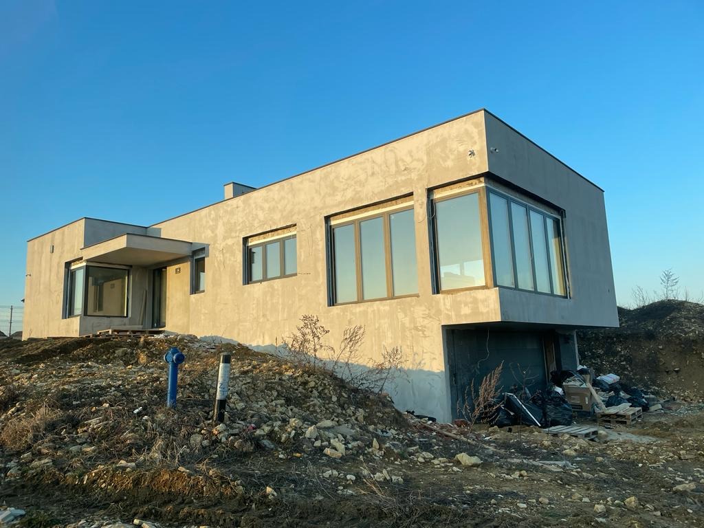 Nowoczesny dom w trakcie budowy, z szarymi oknami, surową elewacją i stertą gruzu przed budynkiem w słoneczny dzień.