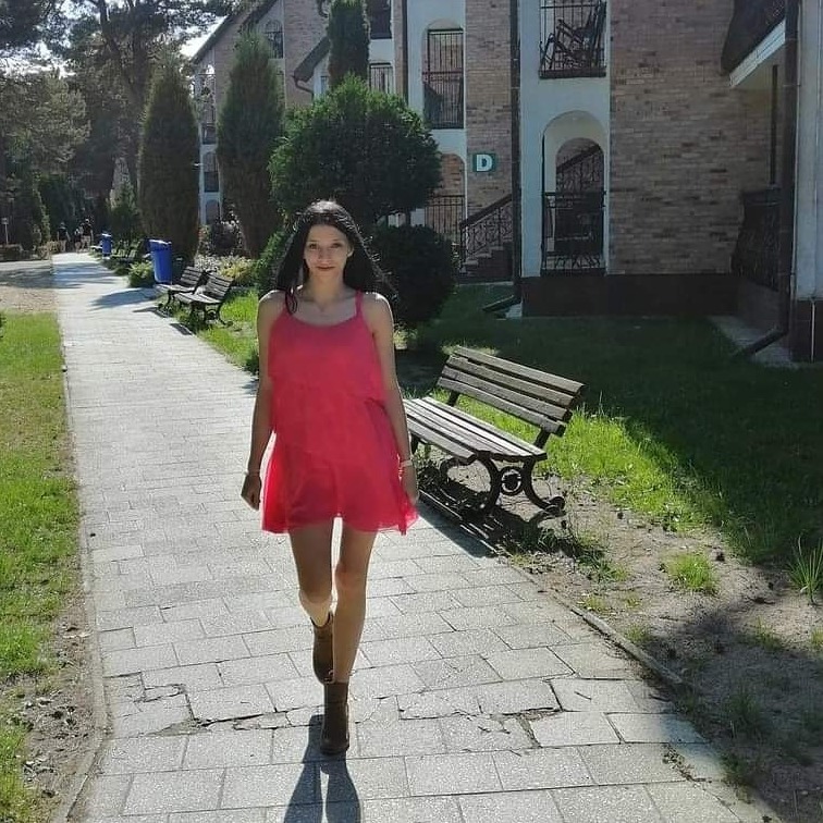 Młoda kobieta w zwiewnej, różowej sukience i brązowych botkach spaceruje po brukowanym chodniku wzdłuż budynku z balkonami i ławek.