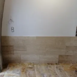 Fragment ściany i podłogi wyłożonej płytkami z trawertynu w trakcie prac wykończeniowych, widoczne wystające kable i puszka elektryczna.