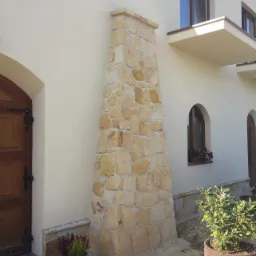 Kamienna obudowa komina zewnętrznego z piaskowca, dopasowana kolorystycznie do elewacji domu jednorodzinnego z drewnianą stolarką okienną i łukowym wejściem.