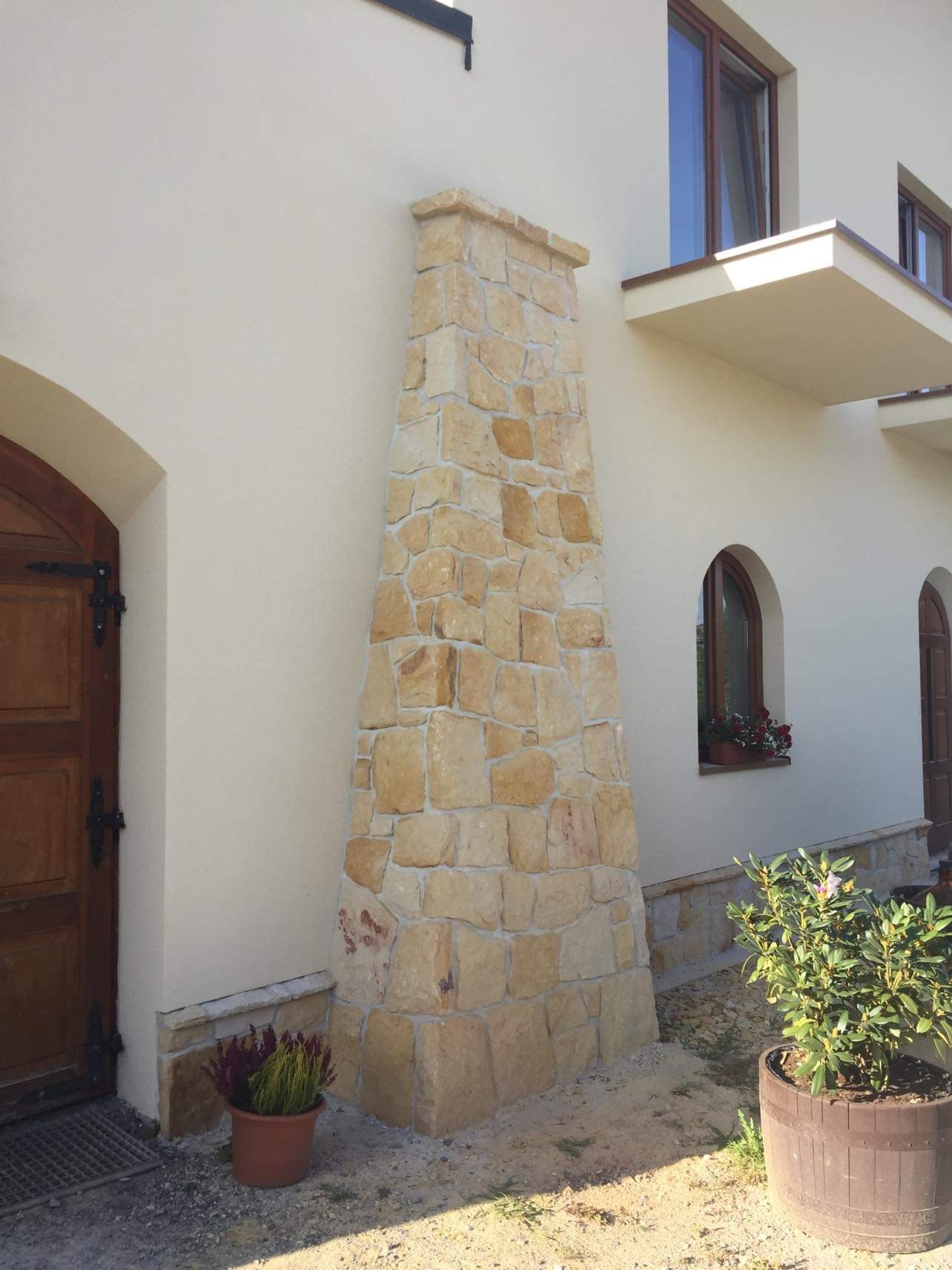 Kamienna obudowa komina zewnętrznego z piaskowca, dopasowana kolorystycznie do elewacji domu jednorodzinnego z drewnianą stolarką okienną i łukowym wejściem.