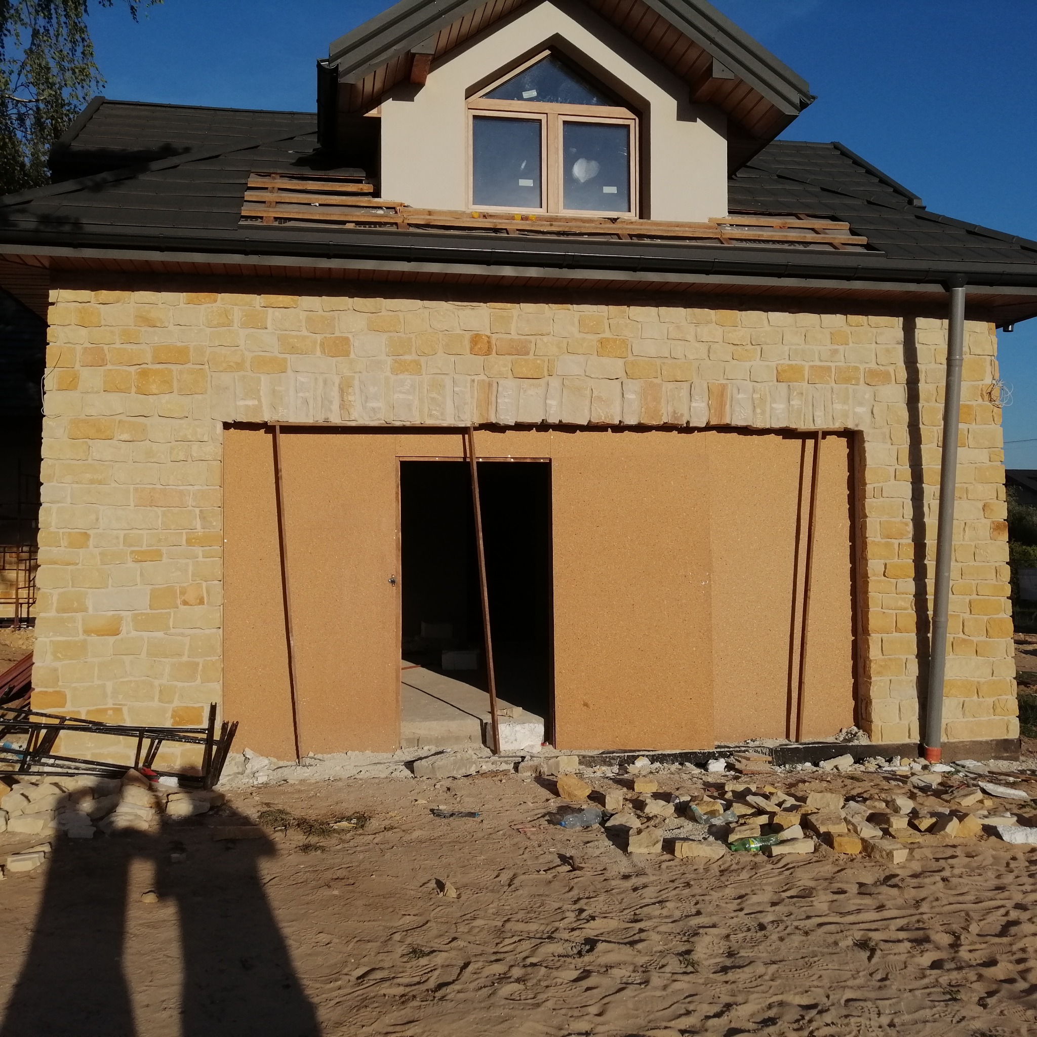 Wznoszony budynek z żółtej cegły, z czarnym dachem i oknem na poddaszu, zabezpieczony płytami OSB zamiast bramy garażowej, z widocznym rusztowaniem i gruzem na ziemi.