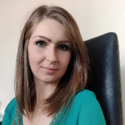 Portret kobiety z brązowymi włosami i zielonymi oczami, ubrana w turkusową bluzkę z ozdobnym dekoltem, siedzącej na czarnym skórzanym krześle.