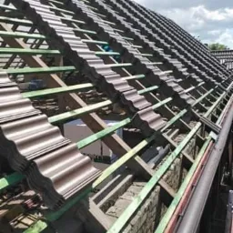 Montaż brązowej dachówki falistej na drewnianej konstrukcji więźby dachowej, widok z boku na częściowo pokryty dach, z zamontowaną rynną w kolorze brązowym.