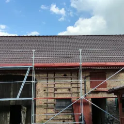 Nowy, brązowy dach z dachówki ceramicznej na budynku gospodarczym z cegły, widoczne rusztowania oraz fragment białego samochodu dostawczego.