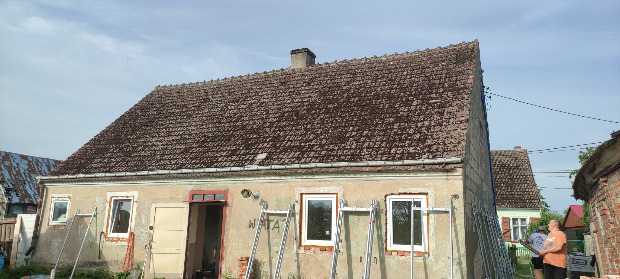 Dom jednorodzinny z widocznym dachem pokrytym starą dachówką porośniętą mchem, z rynnami i kominem, przed budynkiem ustawione drabiny, w tle inne budynki.