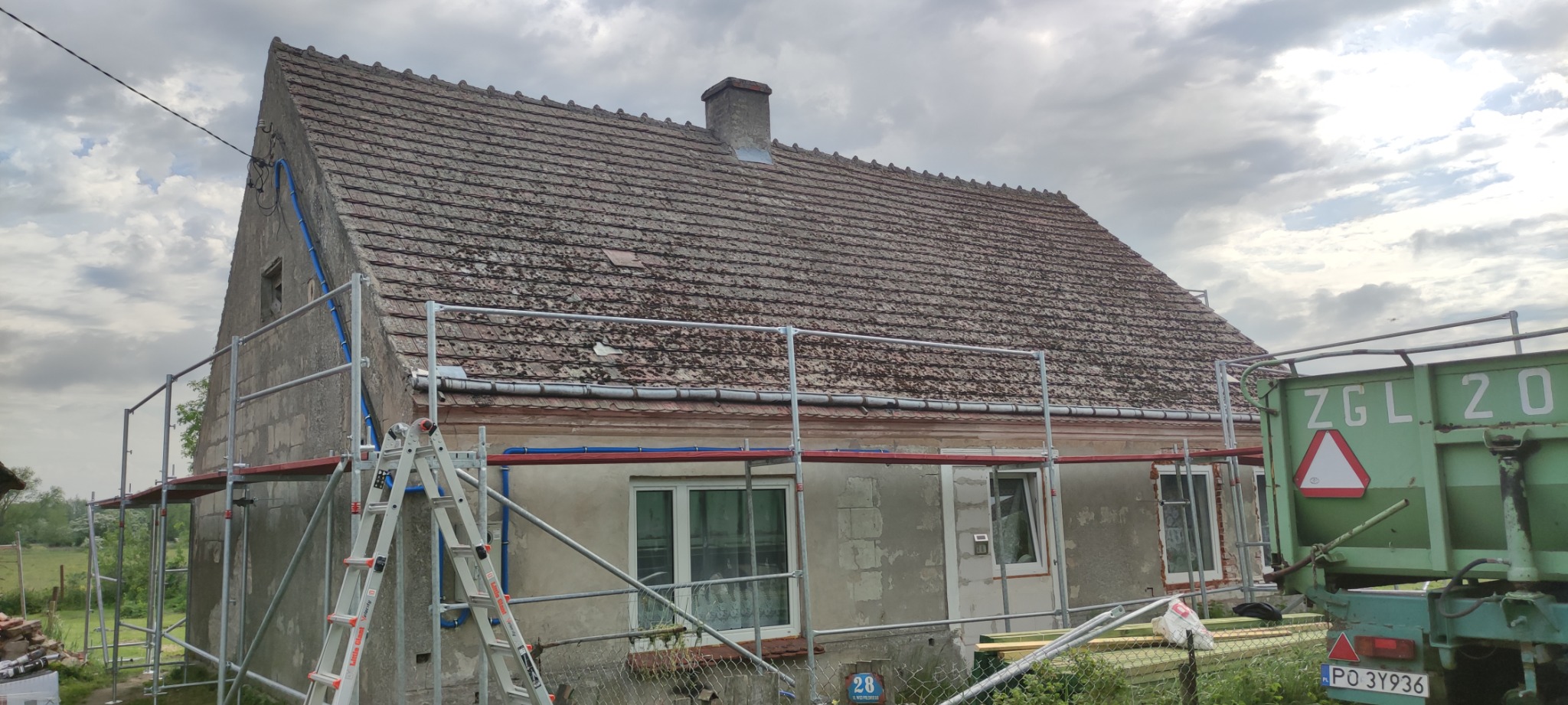 Dom jednorodzinny w trakcie remontu elewacji i dachu, widoczne rusztowania, stara dachówka pokryta mchem, przyczepa z gruzem i drabina oparta o ścianę.