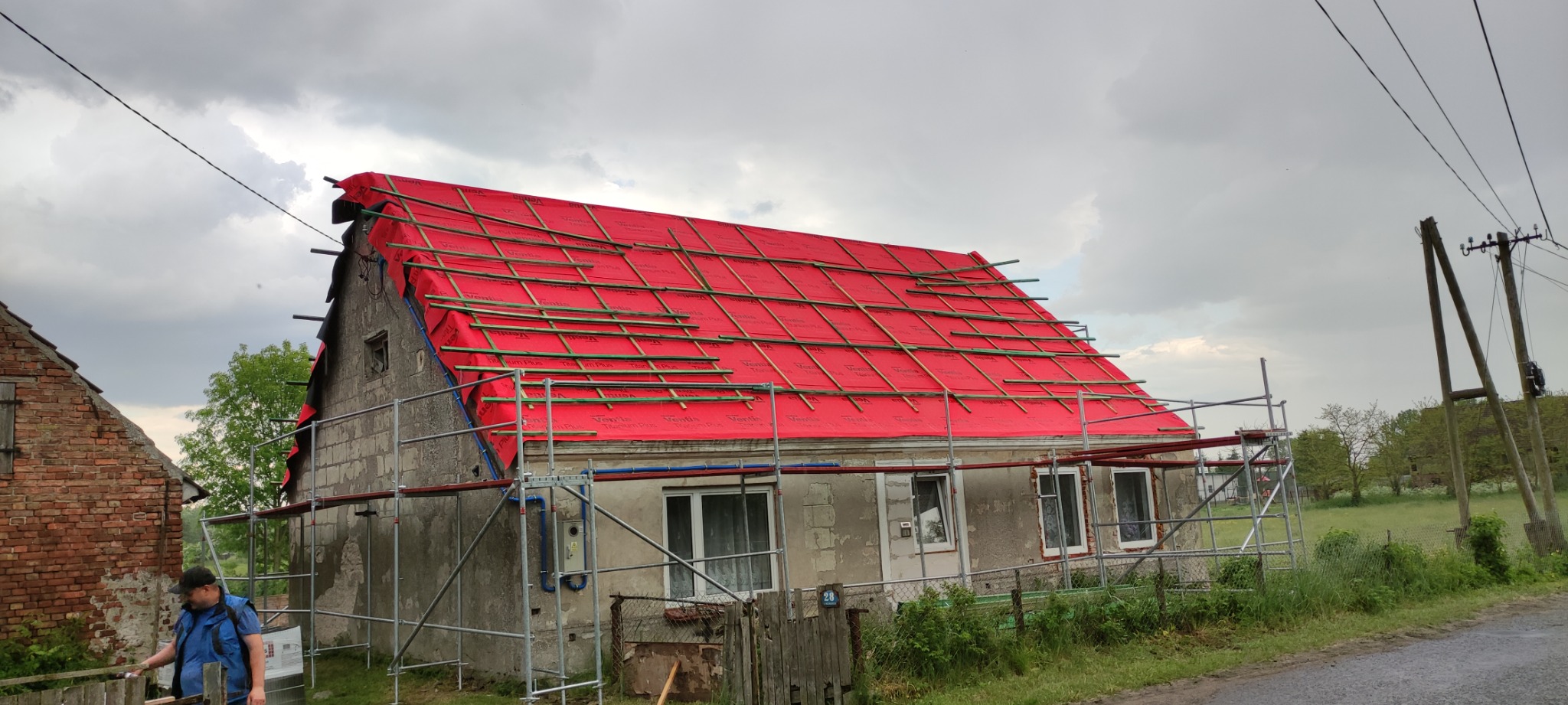 Dom jednorodzinny z częściowo zdjętym starym pokryciem dachowym, zabezpieczony czerwoną membraną i drewnianą łatą, widoczne rusztowania i pracownik w pobliżu.
