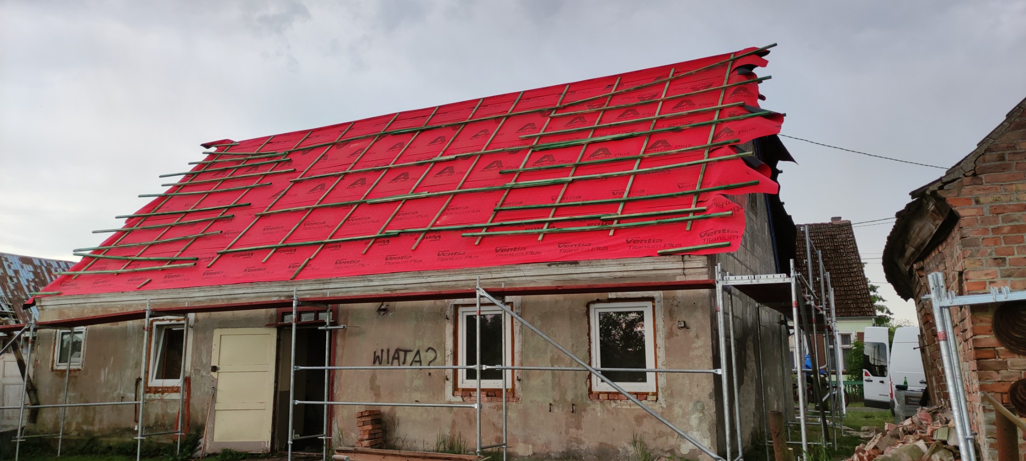 Czerwona membrana dachowa z łatami na budynku w trakcie wymiany dachu, widoczne rusztowania i otoczenie wiejskie.