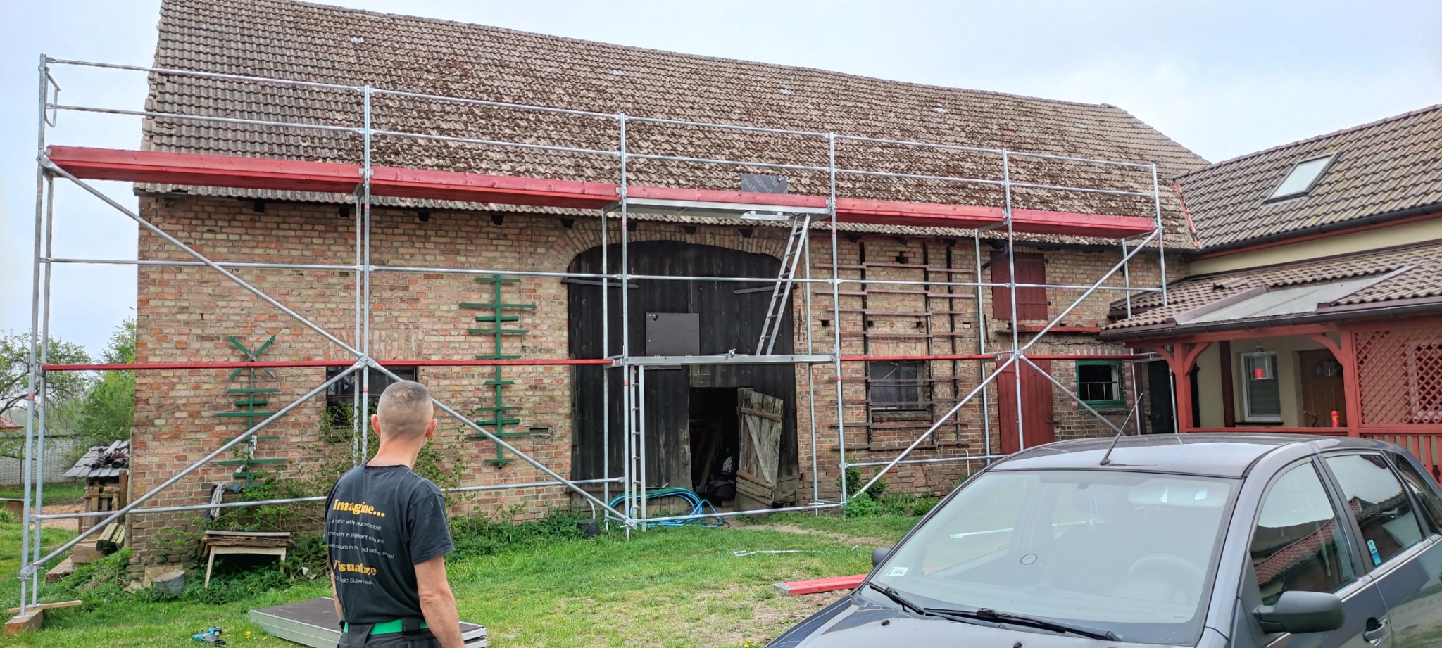 Rusztowanie przy ceglanym budynku gospodarczym z dachówką, widoczny mężczyzna w koszulce z napisem i fragment samochodu