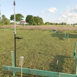 Pomiar geodezyjny na łące z użyciem niwelatora, widoczne drewniane znaczniki. W tle dom i drzewa pod błękitnym niebem.